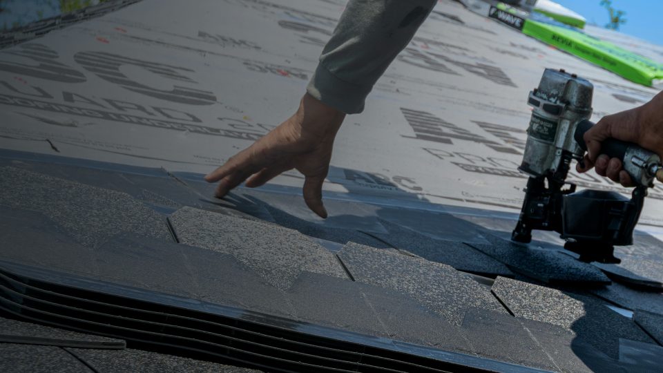 A detailed close-up showing a contractor's hands placing dark asphalt shingles over printed roofing underlayment and securing them with a coil nail gun.
